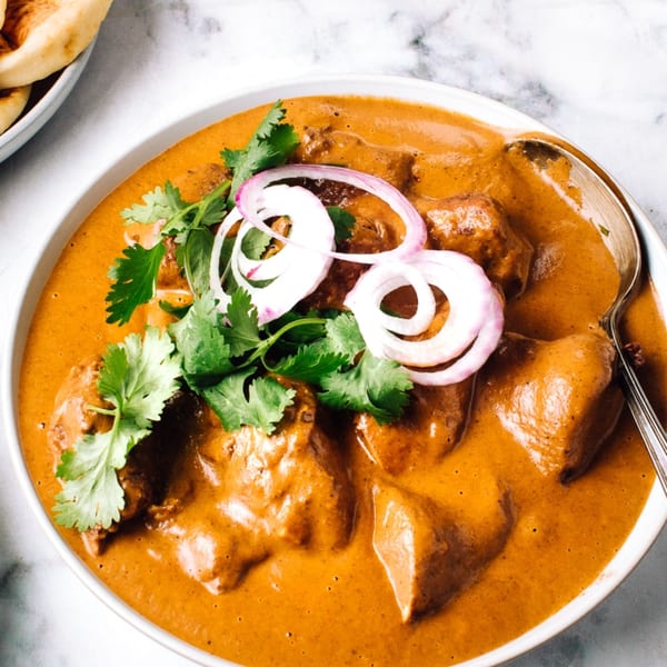 A bowl of Indian butter chicken, garnished with red onion rings and fresh cilantro, with naan bread.