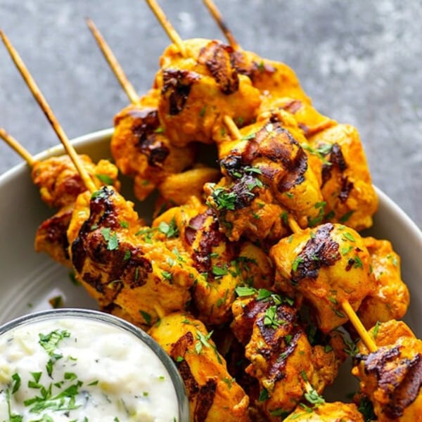 Indian chicken tikka skewers with charred edges and fresh parsley, served with a side of raita.