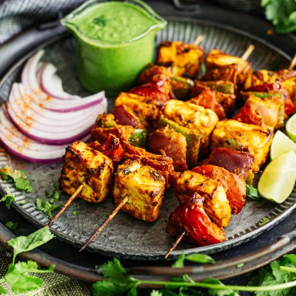 Indian paneer tikka skewers with bell peppers, onions, lime, and green chutney on a metal plate.