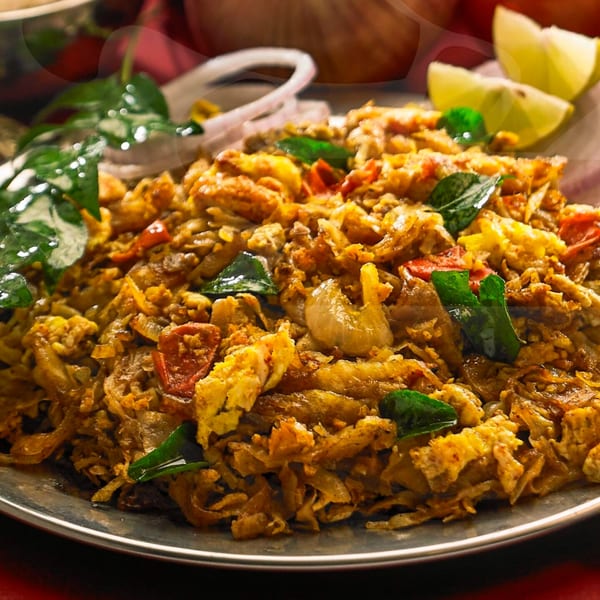 Indian Kothu Parotta with chicken, egg, vegetables, curry leaves, and lime wedges on a metal plate.